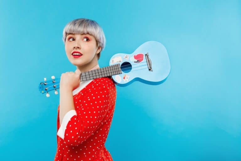 Close up portrait of beautiful dollish girl with short light violet hair wearing red dress holding ukulele over blue background.